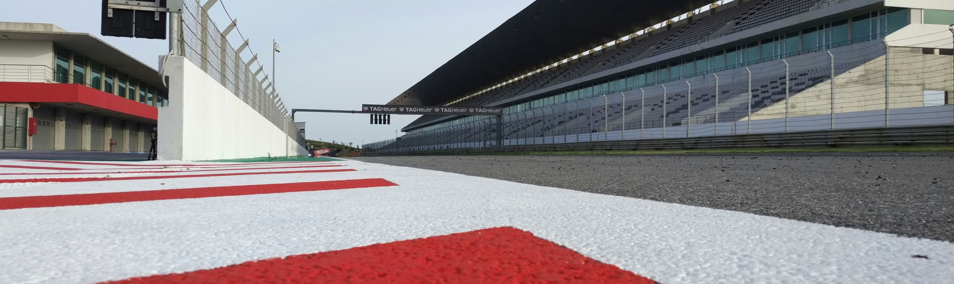 Vue d'une piste de course avec des lignes rouges et blanches, des tribunes modernes à droite, et un ciel clair.