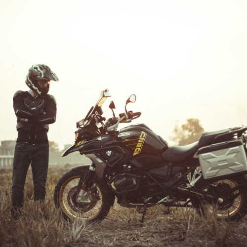 Un motard debout à côté d'une moto dans un champ, portant un casque et une tenue de moto, avec un ciel clair en arrière-plan.